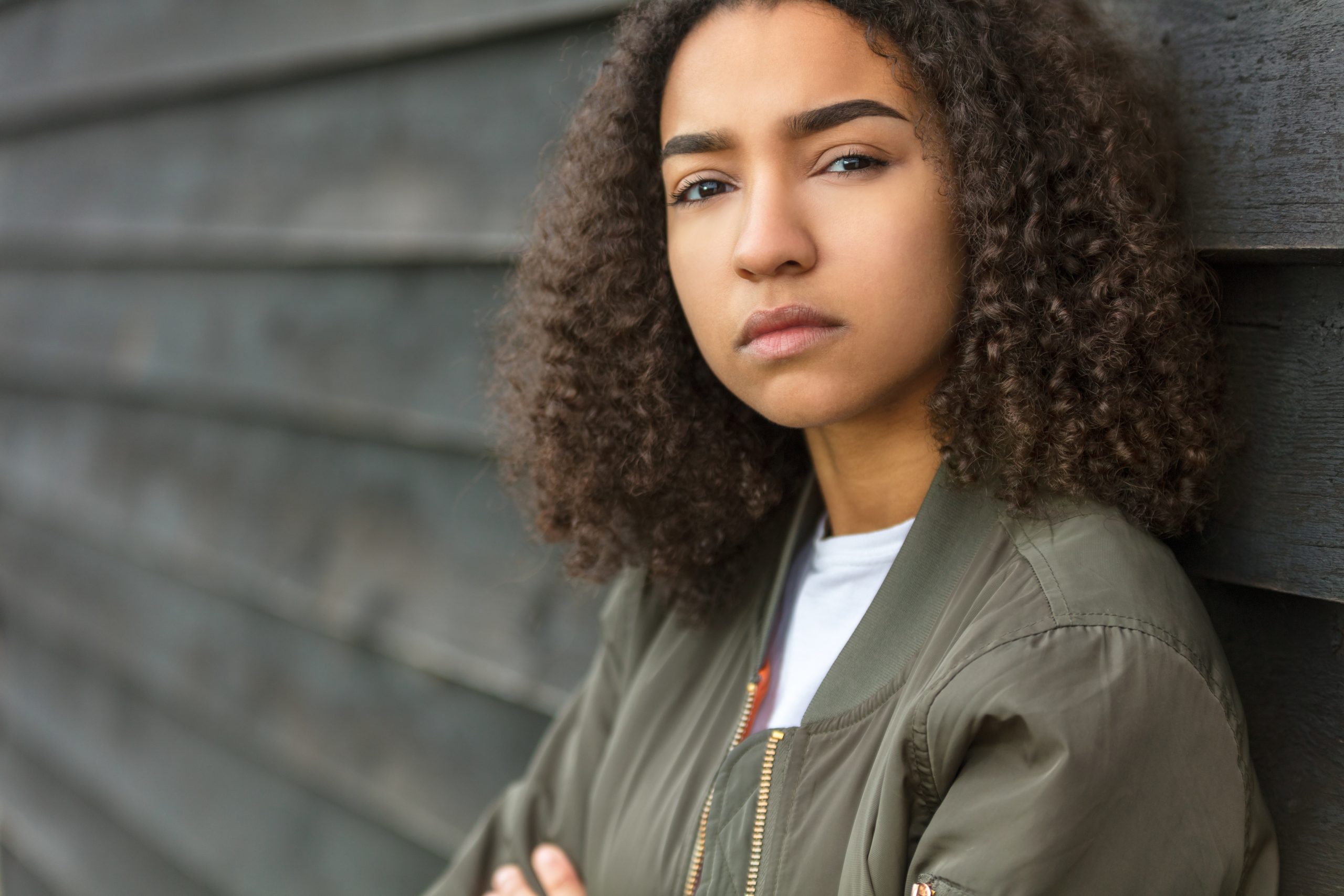 Beautiful mixed race African American girl teenager female young woman outside wearing a green bomber jacket looking sad depressed or thoughtful
