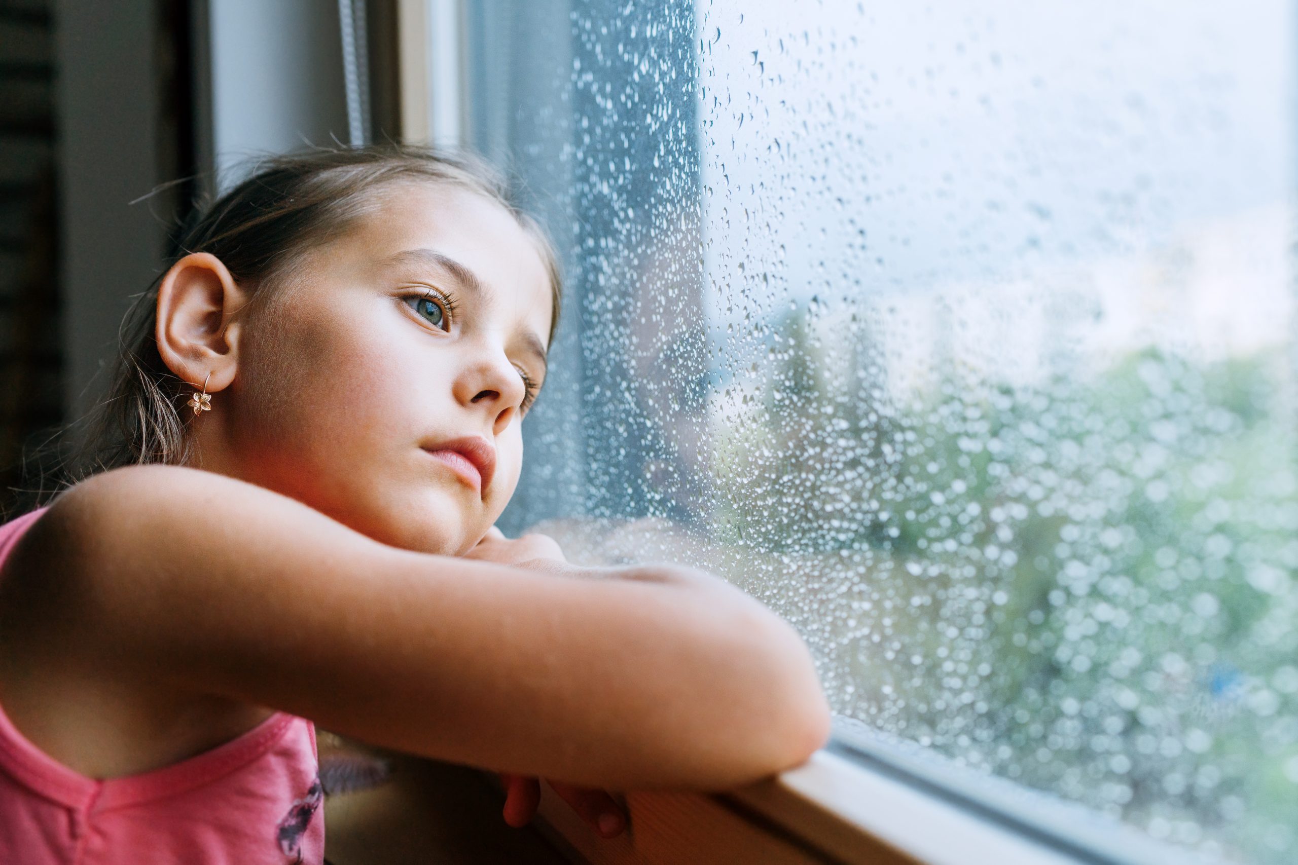 Little sad girl pensive looking through the window glass with a lot of raindrops.